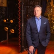 Professional headshot of a person wearing a blue blazer and light blue dress shirt, facing the camera against a softly lit indoor background with warm tones and decorative architectural details. The portrait is framed from the chest up, with a patterned pocket square visible in the jacket pocket.