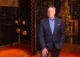 Professional headshot of a person wearing a blue blazer and light blue dress shirt, facing the camera against a softly lit indoor background with warm tones and decorative architectural details. The portrait is framed from the chest up, with a patterned pocket square visible in the jacket pocket.