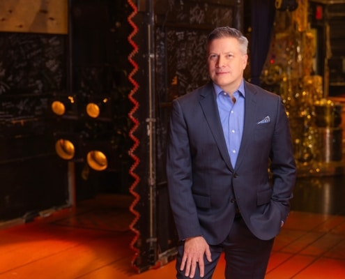 Professional headshot of a person wearing a blue blazer and light blue dress shirt, facing the camera against a softly lit indoor background with warm tones and decorative architectural details. The portrait is framed from the chest up, with a patterned pocket square visible in the jacket pocket.