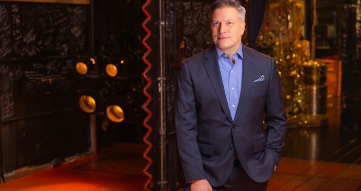 Professional headshot of a person wearing a blue blazer and light blue dress shirt, facing the camera against a softly lit indoor background with warm tones and decorative architectural details. The portrait is framed from the chest up, with a patterned pocket square visible in the jacket pocket.