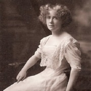 Black-and-white portrait of a seated person in an early 20th-century lace dress, photographed in a studio setting with a softly blurred background.
