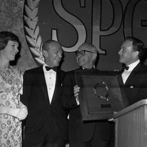 Black-and-white photograph of four people in formal evening attire standing on a stage at an awards ceremony. One person at center holds a large framed plaque featuring a laurel wreath design while another stands at a podium. Large letters forming part of a backdrop are visible behind the group. The image appears to document a formal recognition or industry event.
