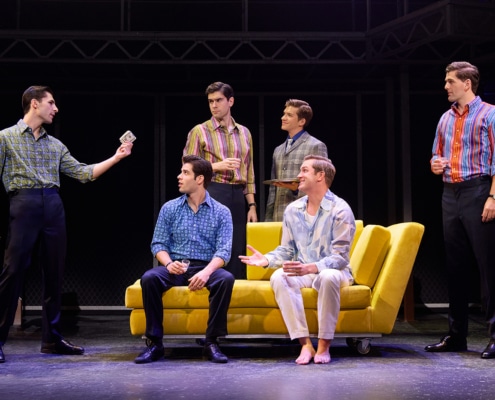 Six performers gather around a bright yellow couch onstage in a scene from Jersey Boys. One performer stands holding a dollar bill while others sit or stand nearby in colorful patterned shirts, with an industrial-style set and staircases in the background.