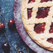 Classic cherry pie with a golden lattice crust and bright red cherry filling, surrounded by fresh cherries on a dark, flour-dusted surface.