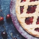Classic cherry pie with a golden lattice crust and bright red cherry filling, surrounded by fresh cherries on a dark, flour-dusted surface.
