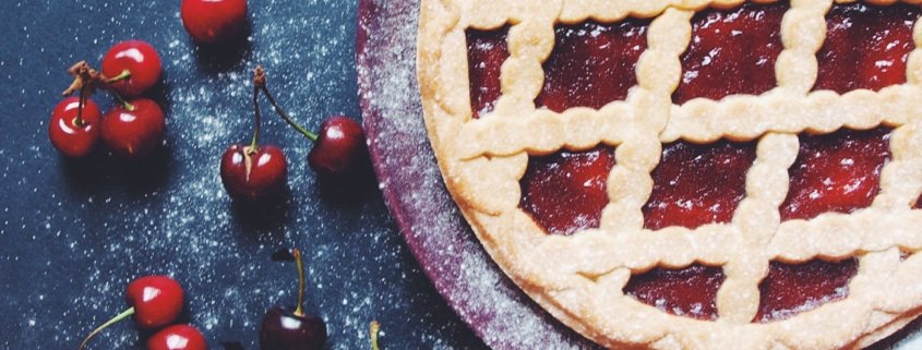 Classic cherry pie with a golden lattice crust and bright red cherry filling, surrounded by fresh cherries on a dark, flour-dusted surface.