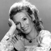 Black-and-white portrait of a person with hair styled up, resting one elbow on a surface and one hand against the side of the head, wearing a patterned long-sleeve top and pendant necklace.