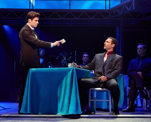 Two performers face each other across a table draped in a blue tablecloth during a scene from Jersey Boys. One performer extends a dollar bill toward the seated performer while other cast members and musicians are visible in the dimly lit background.