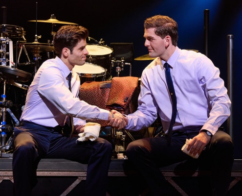 Two performers seated on a stage platform shake hands in front of a drum set during a scene from Jersey Boys. Both wear dress shirts and ties, with stage lighting illuminating the musicians’ equipment in the background.