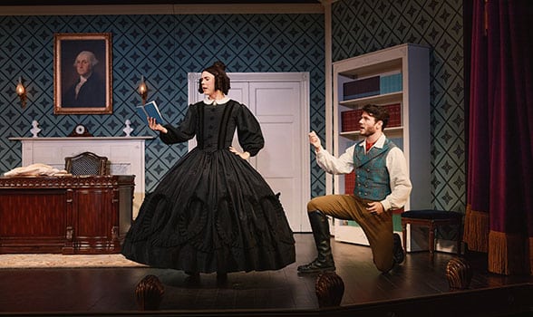 Two performers in period costume stand on a parlor set stage, with one dressed in a full black 19th-century gown gesturing center stage while another kneels beside a bookcase. A desk, wallpapered walls, and a portrait complete the historic interior setting from Oh, Mary!.