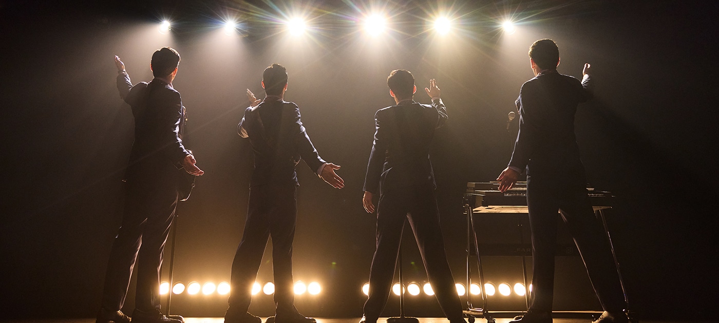 Four performers stand onstage with their backs to the audience beneath bright concert lights in a scene from Jersey Boys. Dressed in matching dark suits, the performers gesture toward the audience as microphones and musical equipment are visible onstage.