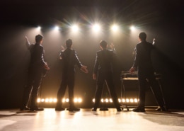 Four performers stand onstage with their backs to the audience beneath bright concert lights in a scene from Jersey Boys. Dressed in matching dark suits, the performers gesture toward the audience as microphones and musical equipment are visible onstage.