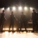 Four performers stand onstage with their backs to the audience beneath bright concert lights in a scene from Jersey Boys. Dressed in matching dark suits, the performers gesture toward the audience as microphones and musical equipment are visible onstage.
