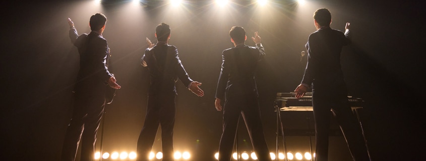 Four performers stand onstage with their backs to the audience beneath bright concert lights in a scene from Jersey Boys. Dressed in matching dark suits, the performers gesture toward the audience as microphones and musical equipment are visible onstage.
