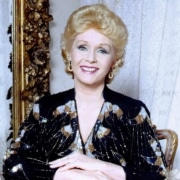 Color portrait of a person seated indoors, wearing a black and gold patterned outfit and earrings, with hands folded and an ornate frame visible in the background.