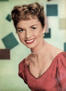 Color studio portrait of actor and singer Debbie Reynolds wearing a rose-colored dress, pearl necklace, and pearl earrings. Reynolds faces the camera against a softly focused background with abstract geometric shapes.
