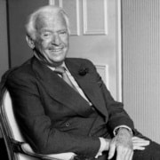 Black-and-white portrait of an older person seated in an upholstered chair, wearing a double-breasted suit and tie, posed indoors in front of decorative wall paneling.