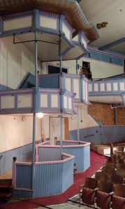 Historic theater interior featuring multiple tiers of pastel blue and pink box seats with decorative woodwork, overlooking rows of audience seating on a red-carpeted floor. Historic theater interior featuring multiple tiers of pastel blue and pink box seats with decorative woodwork, overlooking rows of audience seating on a red-carpeted floor.