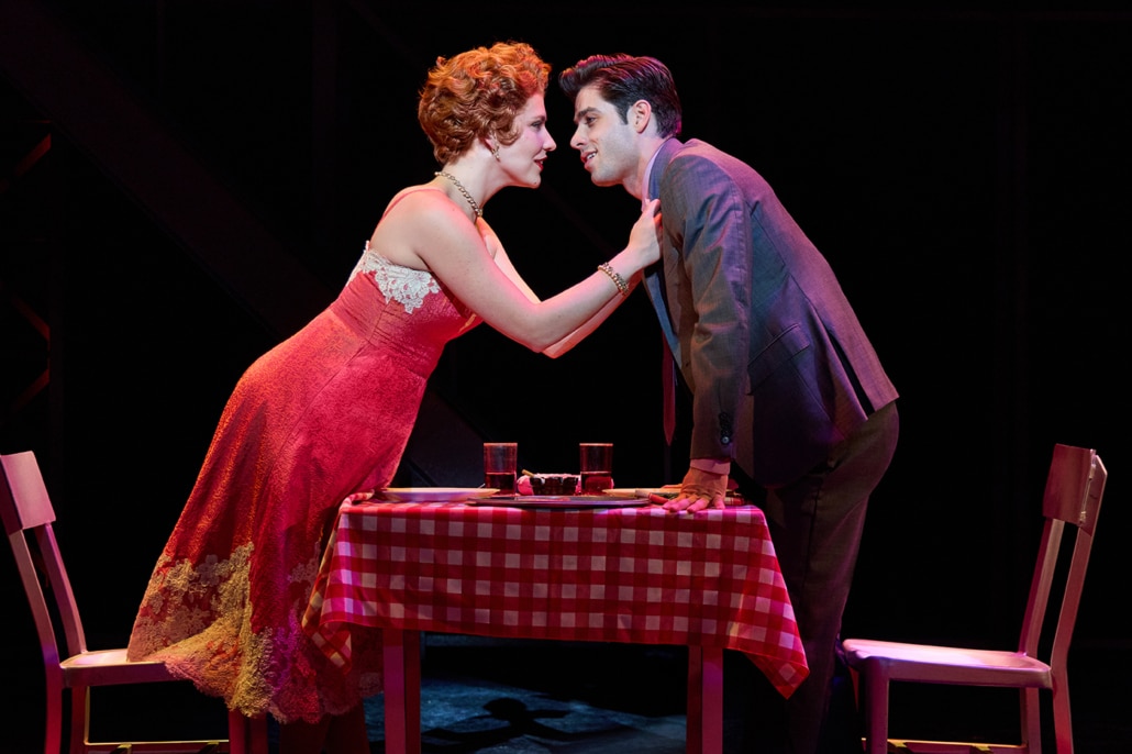 Two performers lean across a small table covered with a red-and-white checkered tablecloth in a scene from Jersey Boys. One performer in a red dress reaches toward another performer in a gray suit, with plates and drinks arranged on the table between them.