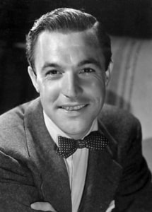 lack-and-white head-and-shoulders studio portrait of Gene Kelly wearing a suit jacket, white shirt, and polka-dot bow tie. Kelly faces the camera with arms folded against a softly blurred background.