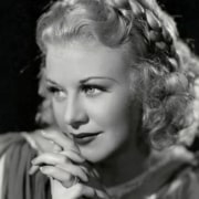 Black-and-white studio portrait showing a person with curled hair resting clasped hands beneath the chin, illuminated by soft lighting against a dark background.