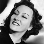 Black-and-white close-up portrait of a person with dark wavy hair looking toward the camera, featuring dramatic lighting and classic Hollywood-era styling.