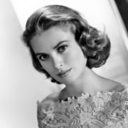Black-and-white studio portrait of a person with short curled hair, wearing a lace-patterned off-the-shoulder gown and facing the camera against a softly lit backdrop.