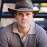 Professional headshot of a person wearing a wide-brimmed gray hat and a light-colored collared knit shirt, seated and facing the camera. The portrait is tightly cropped, with a softly blurred outdoor background featuring dark blue structural elements.