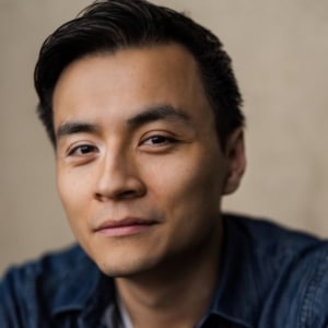 Professional headshot of a person with short dark hair wearing a dark blue shirt, photographed against a neutral beige background. The image is tightly cropped and focuses on the face and upper shoulders.