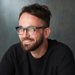 Professional headshot of a person wearing dark-framed glasses and a black sweater, seated against a gray studio backdrop. The portrait is lit with soft natural-looking light and shows the subject turned slightly to one side.