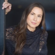Professional headshot of a person with long dark hair wearing a dark patterned top, leaning against a doorway and looking toward the camera. The portrait is softly lit with a dark, out-of-focus background.