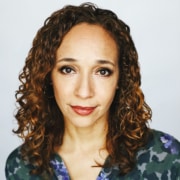 Professional headshot of a person with shoulder-length curly hair, photographed against a light gray background. The person is facing the camera and wearing a patterned green and purple top. The image is tightly cropped from the shoulders up with even studio lighting.