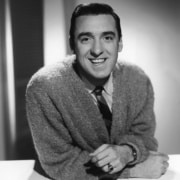 Black-and-white studio portrait of a person in a textured cardigan and tie, leaning forward with folded arms and looking toward the camera against a softly lit background.