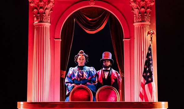 Two performers in period costume sit behind red-upholstered chairs beneath a grand arch framed by columns and draped curtains, with a U.S. flag standing at stage right in a red-lit scene from Oh, Mary!.