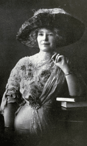 Black-and-white portrait of a person wearing an ornate early 20th-century dress and a wide-brimmed hat, posed beside a small stack of books in a studio setting.