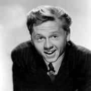 Black-and-white studio portrait of a person in a pinstriped suit and patterned tie, leaning toward the camera with mouth slightly open and hair neatly styled.