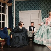Four performers in period costumes gather in a Victorian-style room, including a seated figure in a blue uniform, a figure in a black dress near center stage, a figure holding a metal bucket, and a figure in a mint-green gown. The set includes patterned wallpaper, a tall window, a desk, and a framed portrait from Oh, Mary!.