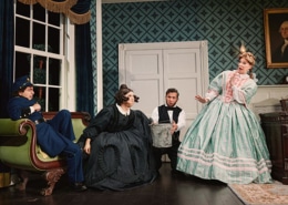 Four performers in period costumes gather in a Victorian-style room, including a seated figure in a blue uniform, a figure in a black dress near center stage, a figure holding a metal bucket, and a figure in a mint-green gown. The set includes patterned wallpaper, a tall window, a desk, and a framed portrait from Oh, Mary!.