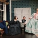 Four performers in period costumes gather in a Victorian-style room, including a seated figure in a blue uniform, a figure in a black dress near center stage, a figure holding a metal bucket, and a figure in a mint-green gown. The set includes patterned wallpaper, a tall window, a desk, and a framed portrait from Oh, Mary!.