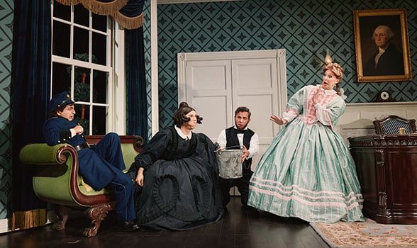 Four performers in period costumes gather in a Victorian-style room, including a seated figure in a blue uniform, a figure in a black dress near center stage, a figure holding a metal bucket, and a figure in a mint-green gown. The set includes patterned wallpaper, a tall window, a desk, and a framed portrait from Oh, Mary!.