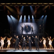 Full company performance scene from Jersey Boys featuring four performers in black suits center stage with raised arms, surrounded by dancers, musicians, and additional cast members beneath dramatic concert lighting and a large illuminated stage structure.