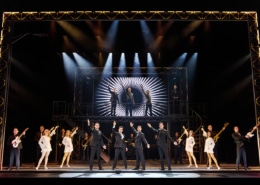 Full company performance scene from Jersey Boys featuring four performers in black suits center stage with raised arms, surrounded by dancers, musicians, and additional cast members beneath dramatic concert lighting and a large illuminated stage structure.