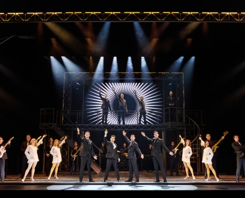 Full company performance scene from Jersey Boys featuring four performers in black suits center stage with raised arms, surrounded by dancers, musicians, and additional cast members beneath dramatic concert lighting and a large illuminated stage structure.
