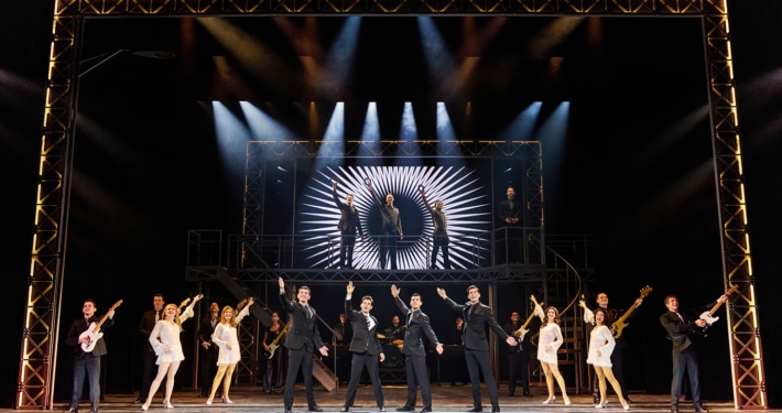 Full company performance scene from Jersey Boys featuring four performers in black suits center stage with raised arms, surrounded by dancers, musicians, and additional cast members beneath dramatic concert lighting and a large illuminated stage structure.