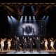 Full company performance scene from Jersey Boys featuring four performers in black suits center stage with raised arms, surrounded by dancers, musicians, and additional cast members beneath dramatic concert lighting and a large illuminated stage structure.