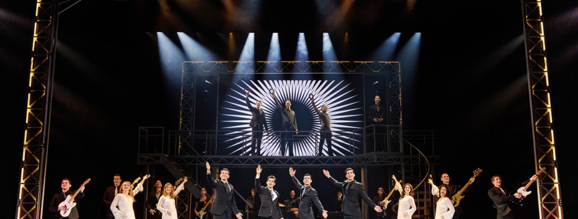 Full company performance scene from Jersey Boys featuring four performers in black suits center stage with raised arms, surrounded by dancers, musicians, and additional cast members beneath dramatic concert lighting and a large illuminated stage structure.
