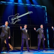Four performers in matching dark suits stand at microphones onstage in a scene from Jersey Boys. Blue lighting and a large illuminated guitar outline frame the concert-style performance.