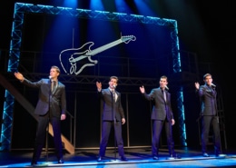 Four performers in matching dark suits stand at microphones onstage in a scene from Jersey Boys. Blue lighting and a large illuminated guitar outline frame the concert-style performance.