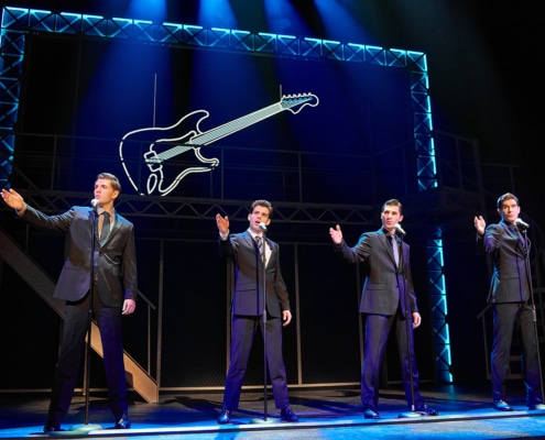 Four performers in matching dark suits stand at microphones onstage in a scene from Jersey Boys. Blue lighting and a large illuminated guitar outline frame the concert-style performance.