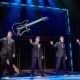 Four performers in matching dark suits stand at microphones onstage in a scene from Jersey Boys. Blue lighting and a large illuminated guitar outline frame the concert-style performance.